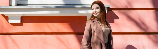 Glückliche rothaarige Frau im Mantel, die auf der Straße neben dem Gebäude steht, Transparent — Stockfoto