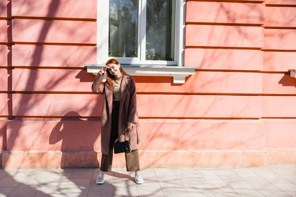 Full length of cheerful redhead woman in coat standing on street near building — стоковое фото