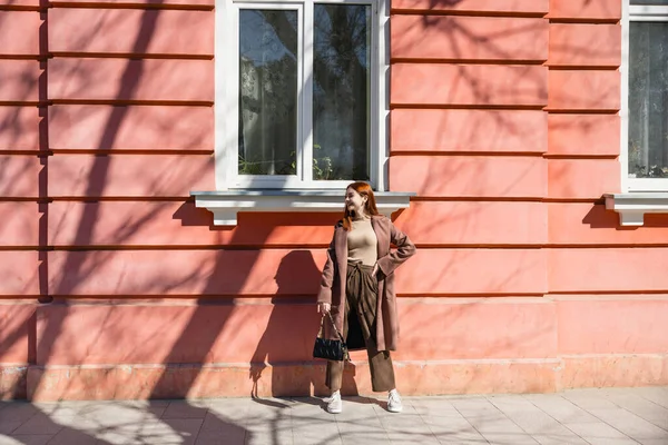 Full length of cheerful redhead woman in coat standing with handbag near building — стоковое фото