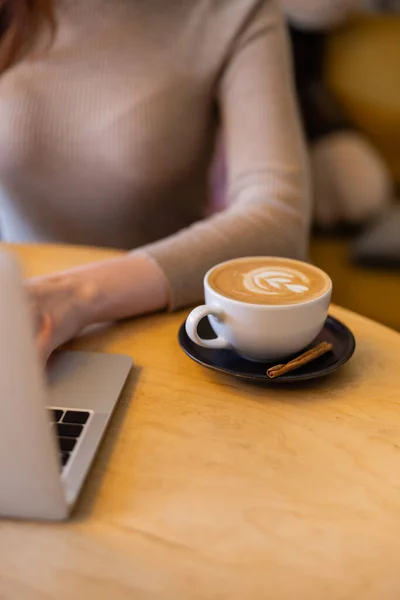 Cropped view of young woman using laptop near cup of cappuccino — Stock Photo