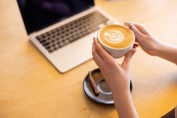 Vista recortada del joven freelancer sosteniendo taza de capuchino cerca de la computadora portátil en la cafetería - foto de stock