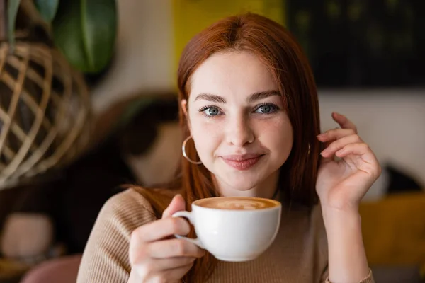 Jeune rousse femme souriant et tenant tasse de latte dans le café — Photo de stock