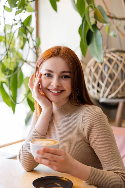 Joven mujer sonriendo y sosteniendo taza de capuchino en la cafetería - foto de stock
