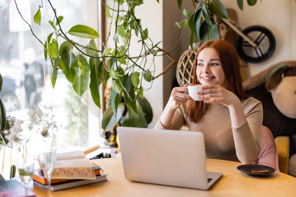 Mujer sonriente con el pelo rojo sosteniendo taza de café cerca de la computadora portátil en la cafetería - foto de stock