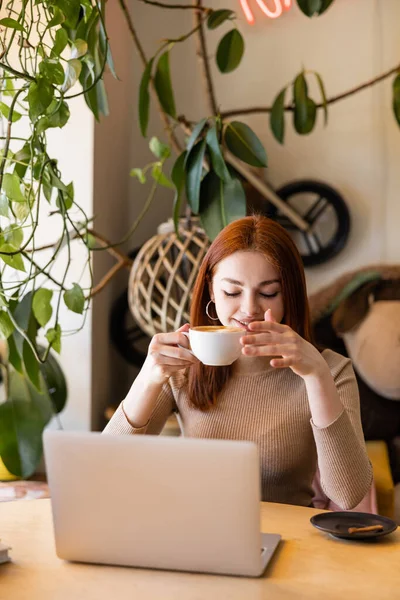 Fröhliche junge Frau mit roten Haaren hält Tasse Kaffee in der Nähe von Laptop im Café — Stockfoto