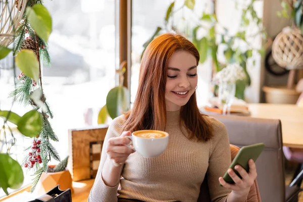 Felice giovane donna con i capelli rossi utilizzando smartphone e tenendo in mano una tazza di caffè nel caffè — Foto stock