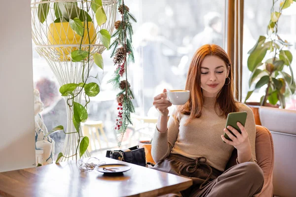 Pleased young woman with red hair using smartphone and holding cup of coffee — Photo de stock