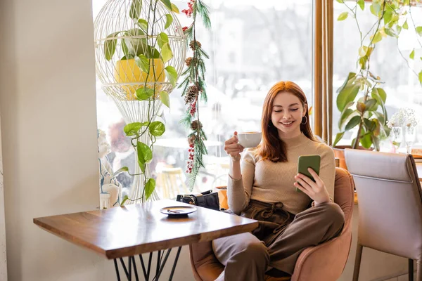 Mujer joven positiva con el pelo rojo usando el teléfono inteligente y sosteniendo la taza de café - foto de stock