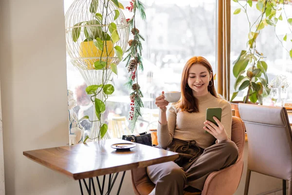 Positive young woman with red hair using smartphone and holding cup of coffee — Stock Photo