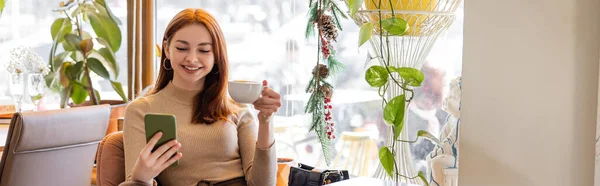 Happy young woman with red hair using smartphone and holding cup of coffee, banner — Stock Photo