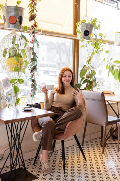 Gioiosa giovane donna con i capelli rossi utilizzando smartphone e tenendo in mano una tazza di caffè sul tavolo — Foto stock