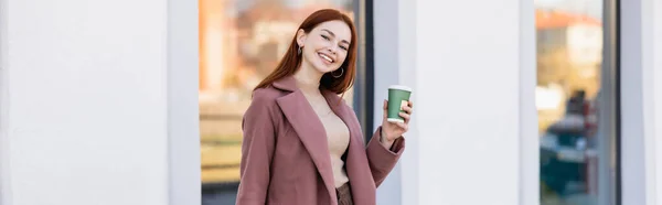 Mujer pelirroja feliz sosteniendo taza de papel en la calle urbana, pancarta - foto de stock