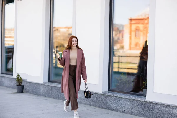 Full length of pleased redhead woman with handbag holding paper cup and walking on urban street — стоковое фото