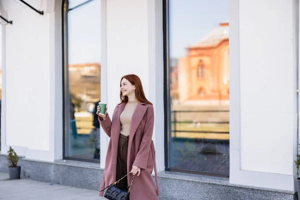 Happy redhead woman with handbag holding paper cup on street — Stock Photo