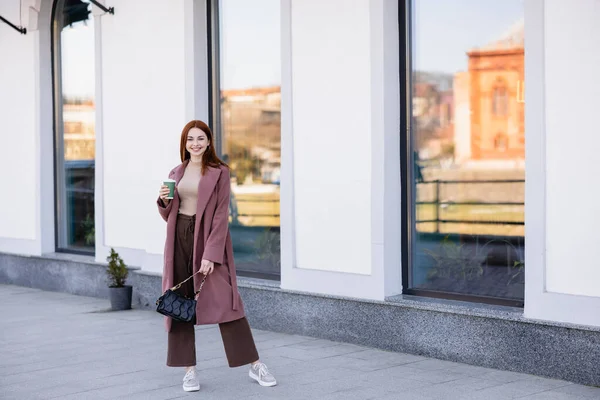 Pleine longueur de femme heureuse avec sac à main tenant tasse en papier sur la rue — Photo de stock