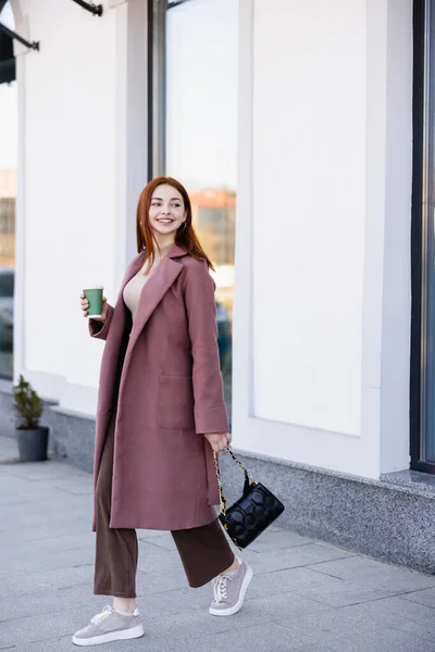 Full length of pleased woman with handbag holding paper cup and walking on urban street — стоковое фото