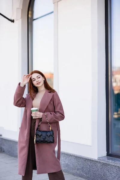 Young woman in coat with handbag adjusting red hair and holding paper cup outside — Photo de stock