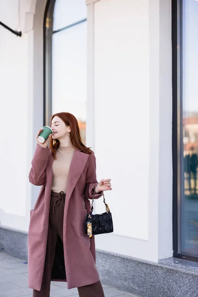 Задоволена жінка з закритими очима п'є каву, щоб піти на міську вулицю — стокове фото