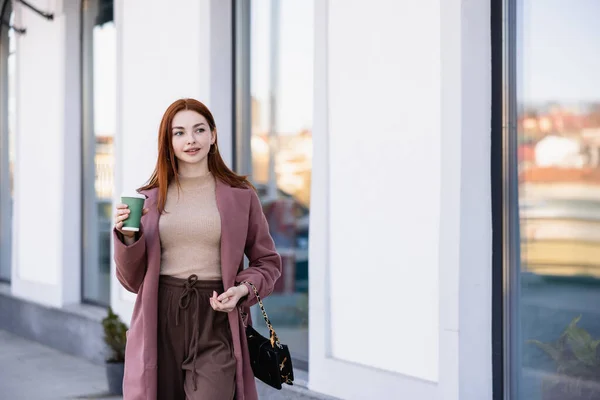 Young pleased woman in coat holding coffee to go on street — Photo de stock