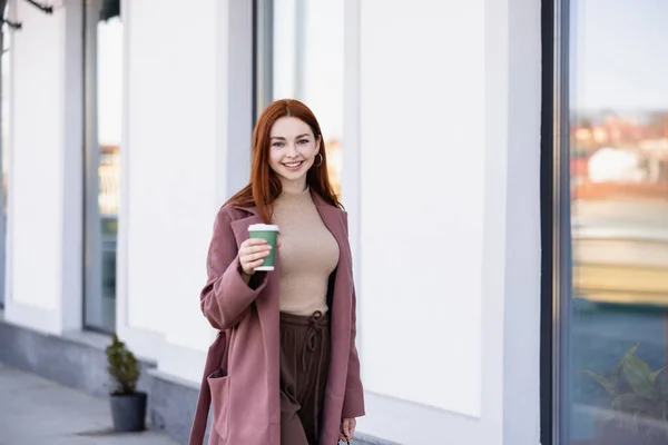 Cheerful woman in coat holding coffee to go on urban street — Photo de stock