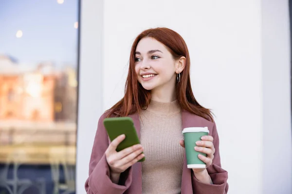 Cheerful woman holding coffee to go and smartphone on street — Photo de stock