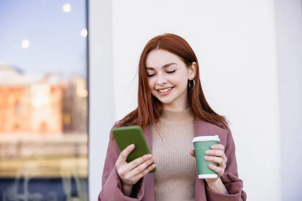 Щаслива жінка тримає каву, щоб піти і смартфон на міській вулиці — стокове фото