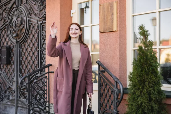 Jeune femme gaie en manteau agitant la main près du bâtiment — Photo de stock