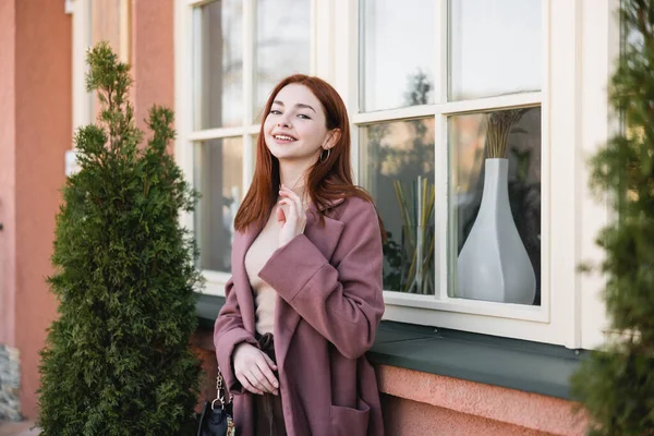 Junge fröhliche Frau im Mantel blickt in die Kamera in der Nähe des Gebäudes — Stockfoto