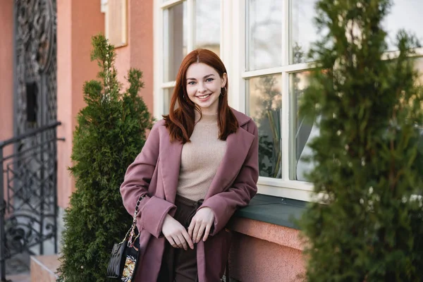 Jeune femme joyeuse aux cheveux roux debout près du bâtiment — Photo de stock