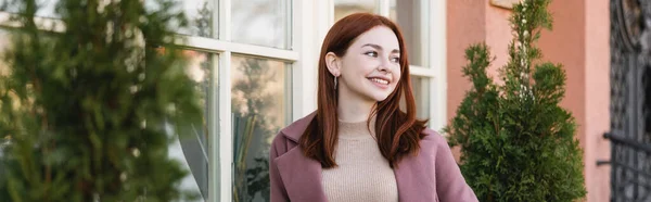 Молода задоволена жінка з рудим волоссям посміхається біля будівлі, банер — стокове фото