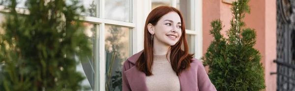 Young pleased woman with red hair smiling near building, banner — Photo de stock