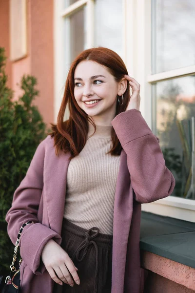 Young pleased woman adjusting red hair near building — стоковое фото