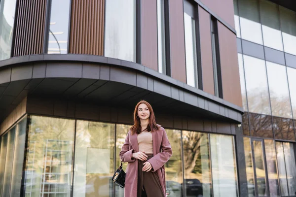 Pleased redhead woman in coat with handbag standing near modern building — стоковое фото