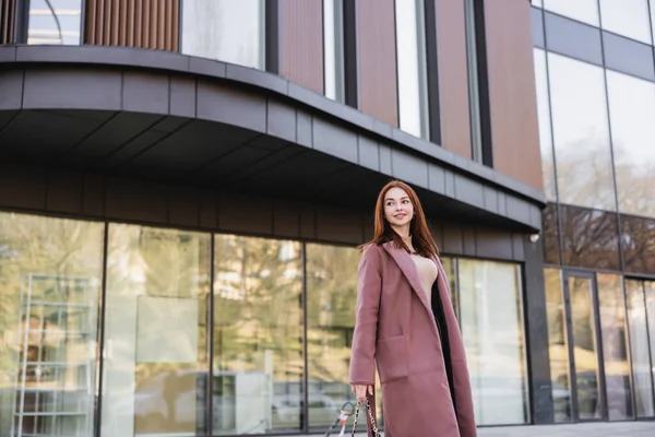 Pleased redhead woman in coat standing near modern building — стоковое фото