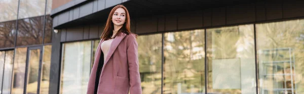 Pleased redhead woman in coat standing near modern building, banner — Stock Photo