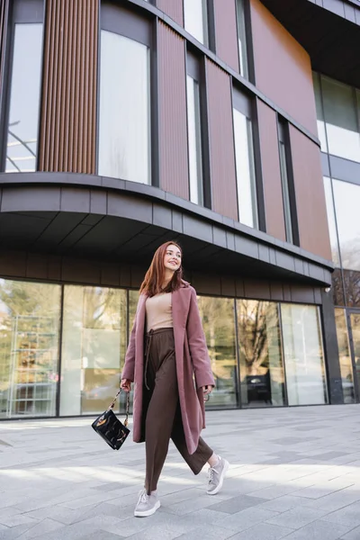 Piena lunghezza di donna rossa soddisfatto in cappotto con borsetta a piedi vicino edificio moderno — Foto stock