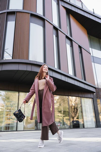 Felice donna rossa in cappotto con borsetta a piedi vicino edificio moderno — Foto stock