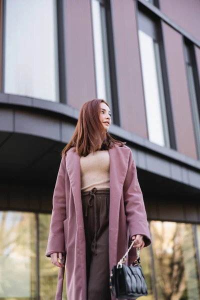 Низький кут зору молодої рудої жінки в пальто з сумкою, що стоїть біля сучасної будівлі — стокове фото