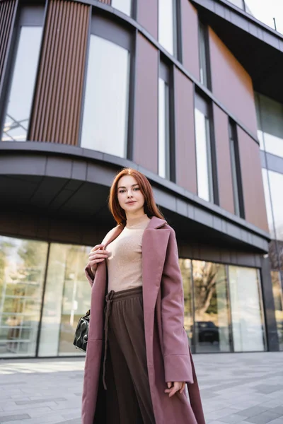 Tiefansicht einer rothaarigen Frau im Mantel, die mit Handtasche in der Nähe moderner Gebäude steht — Stockfoto