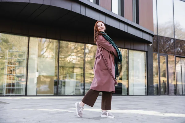 Pleine longueur de femme rousse heureuse en manteau avec sac à main debout près du bâtiment moderne — Photo de stock