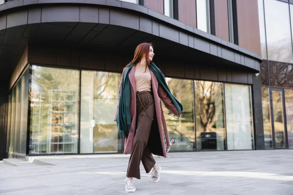 Full length of pleased redhead woman in coat with scarf walking near modern building — Photo de stock
