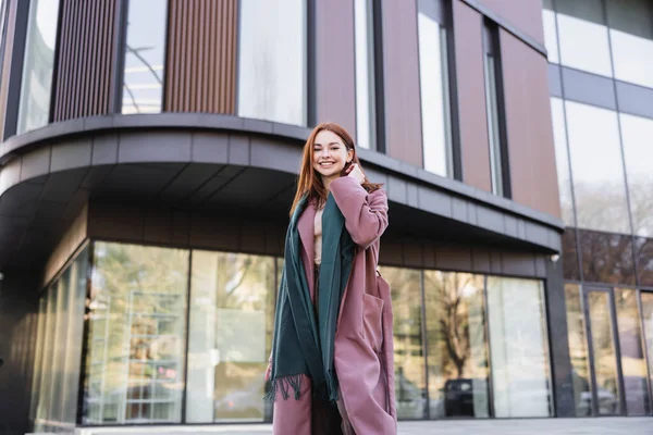 Rothaarige Frau im Mantel mit Schal nahe modernem Gebäude — Stockfoto