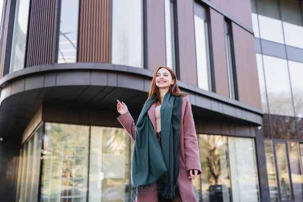 Low angle view of joyful redhead woman in coat with scarf near modern building — стоковое фото