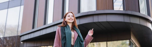 Low angle view of joyful redhead woman in coat with scarf near modern building, banner — Stock Photo