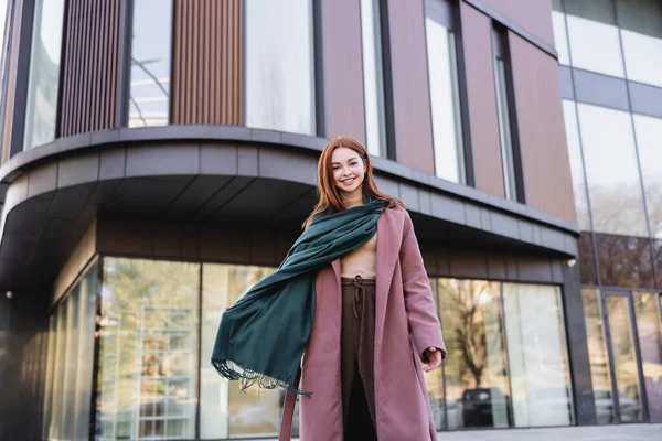 Vue à angle bas de femme rousse heureuse en manteau avec écharpe debout près du bâtiment moderne — Photo de stock