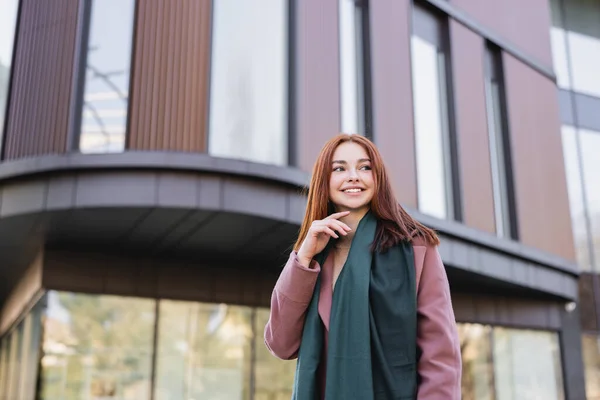 Tiefansicht einer zufriedenen jungen Frau im Mantel mit Schal in der Nähe moderner Gebäude — Stockfoto