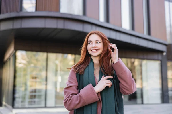 Low angle view of pleased woman in coat with scarf adjusting red hair near modern building - foto de stock