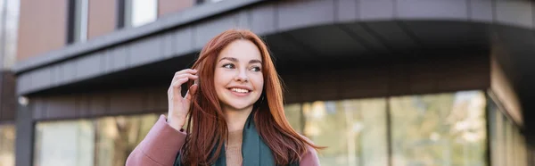 Jovem mulher satisfeita no cachecol ajustando o cabelo vermelho perto do edifício moderno, banner — Fotografia de Stock