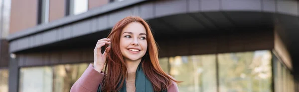 Joven mujer complacida en bufanda ajuste de pelo rojo cerca de edificio moderno, pancarta - foto de stock