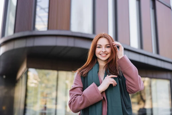 Vista de ángulo bajo de la mujer alegre en abrigo con bufanda de ajuste de pelo rojo cerca de la construcción moderna - foto de stock